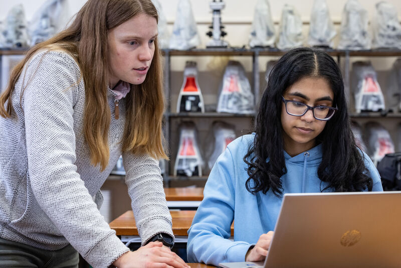 Two students looking at a laptop in a classroom with some microscopes on shelves behind them